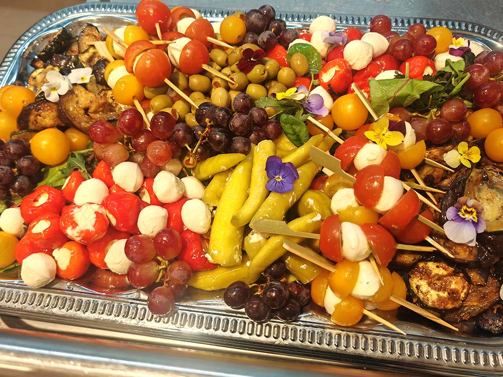 Eine Platte mit farbenfrohen Vorspeisen: Trauben- und Tomatenspieße mit Mozzarella, gefüllte Paprika, Oliven, gegrillte Auberginenscheiben und essbare Blumen, kunstvoll auf einem Silbertablett angerichtet.