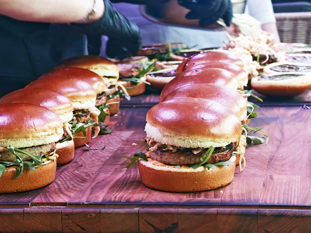 Mehrere große Gourmet-Burger mit Salat und dicken Brötchen sind auf einer Holzfläche aufgereiht, während behandschuhte Hände sie im Hintergrund zubereiten oder zusammensetzen.