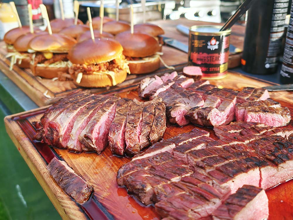 Auf einem hölzernen Schneidebrett sind Scheiben von gegrilltem Steak arrangiert, während im Hintergrund mehrere Pulled-Pork-Sandwiches auf Brötchen liegen. Ein Gewürzglas und Grillutensilien sind ebenfalls zu sehen.
