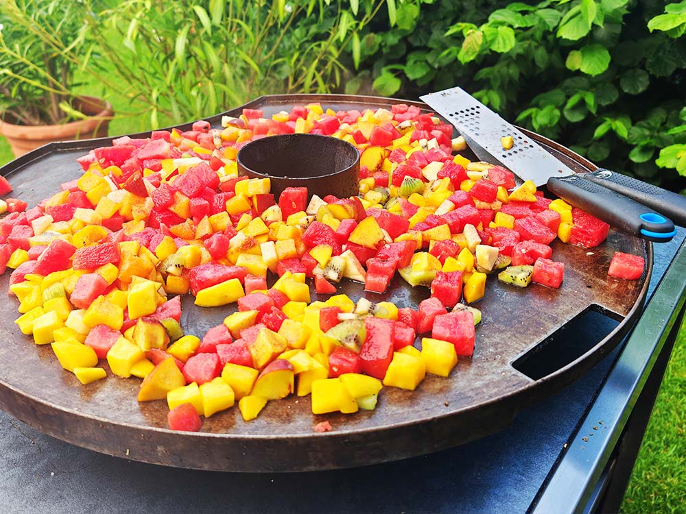 Bunte gewürfelte Früchte, darunter gelbe Mango und rote Wassermelone, liegen auf einem großen runden Grill im Freien mit einer Metallreibe und üppigen Grünpflanzen im Hintergrund.