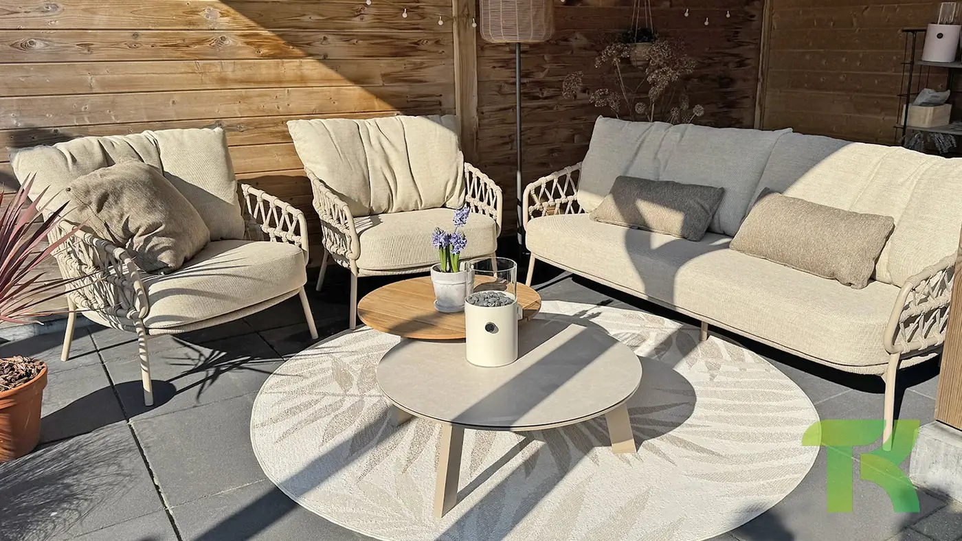 Moderne Terrasse mit beigen gepolsterten Stühlen, Sofa, Emma Couchtisch-Set und Topfpflanzen auf einem runden Teppich.