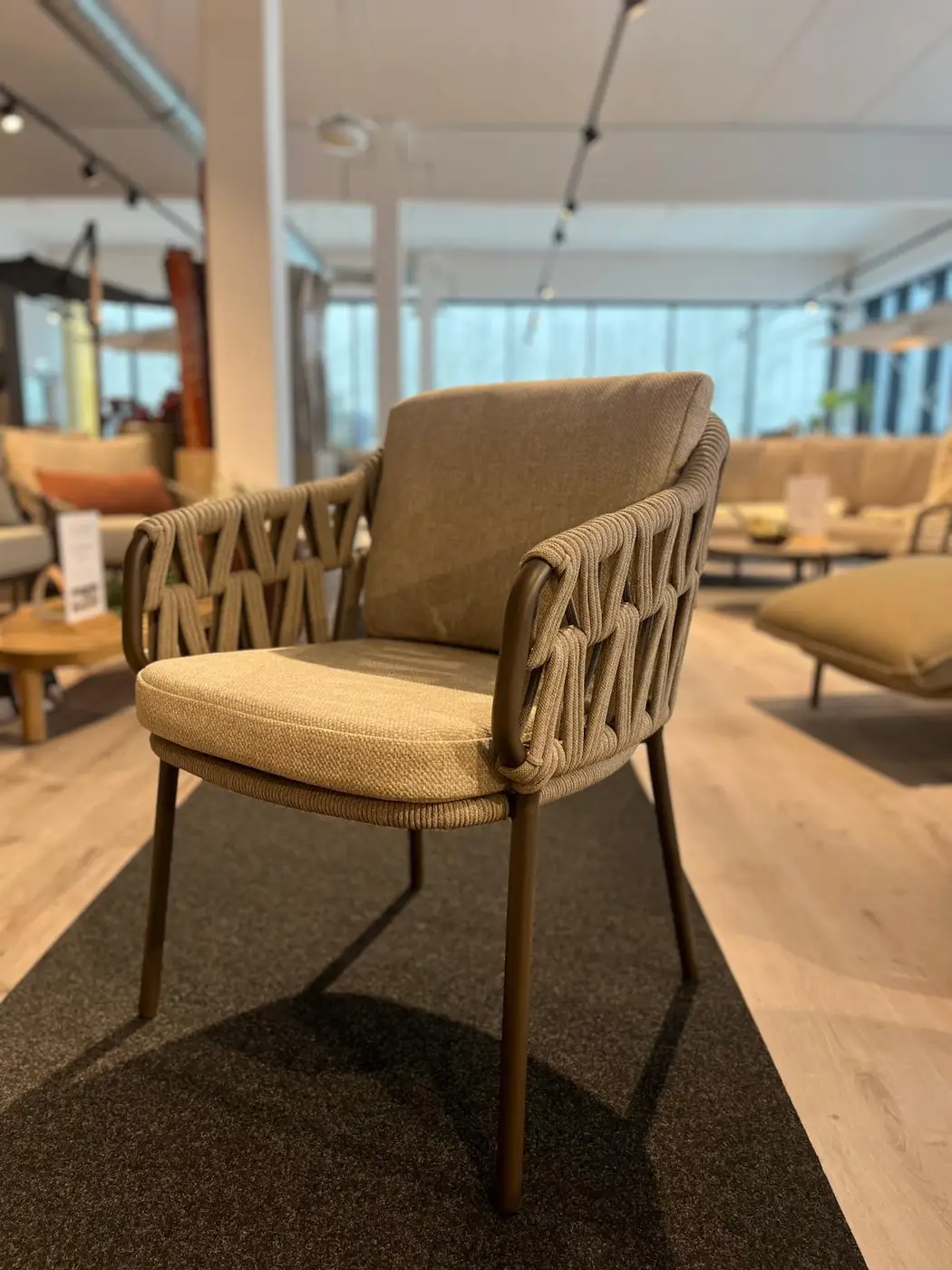 Der Pisa Dining Stuhl ist ein beigefarbener, geflochtener Sessel mit gepolsterter Sitzfläche und Rückenlehne, der in einem modernen Ausstellungsraum gezeigt wird.