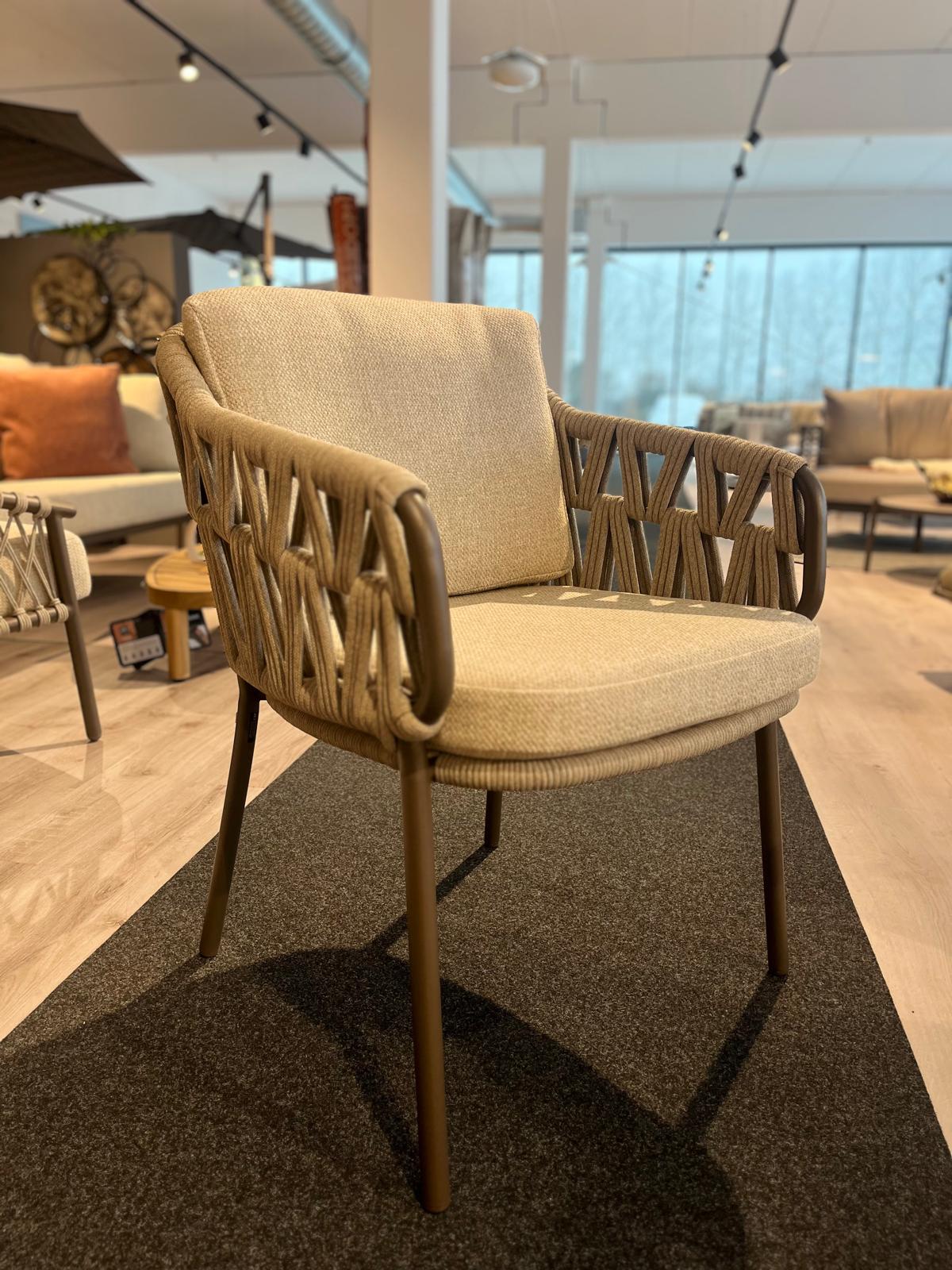 Der Pisa Dining Stuhl, ein beigefarbener gepolsterter Stuhl mit geflochtenen Armlehnen und Metallbeinen, wird in einem modernen Ausstellungsraum präsentiert.