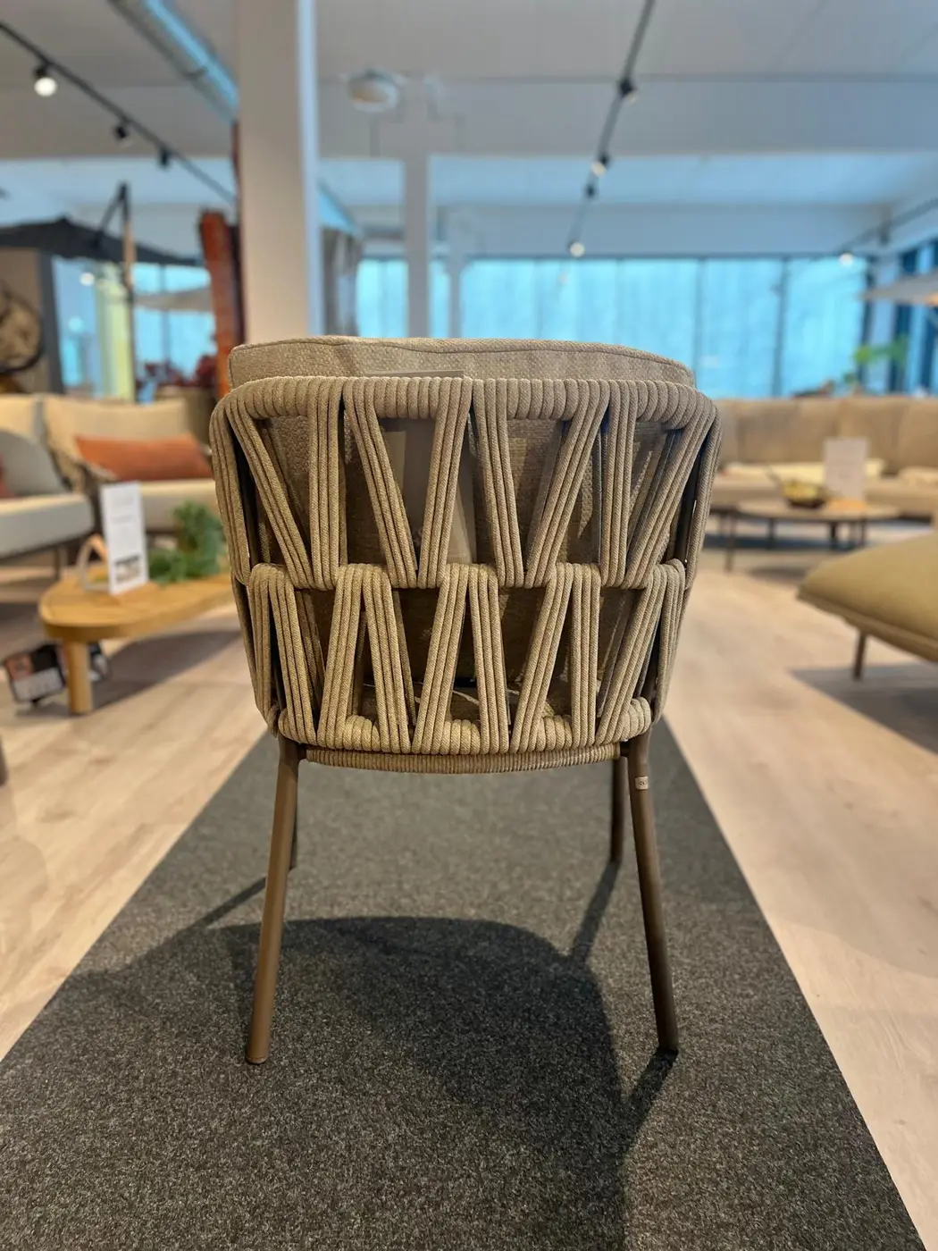 Pisa Dining Stuhl: brauner Flechtstuhl mit Metallbeinen, gezeigt auf einem dunklen Teppich in einem hellen, modernen Ausstellungsraum.