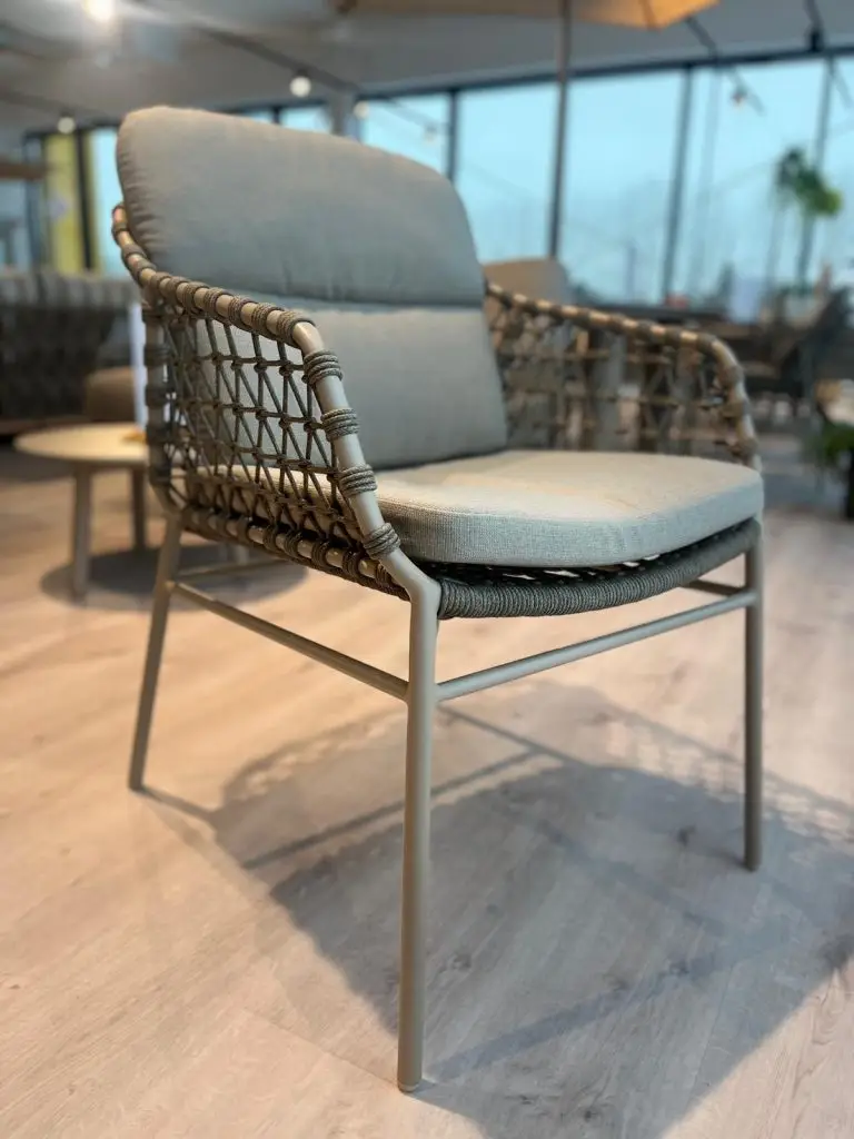 Der Kos Dining Stuhl hat einen gepolsterten Sitz, geflochtene Seiten und Metallbeine auf einem Holzboden.