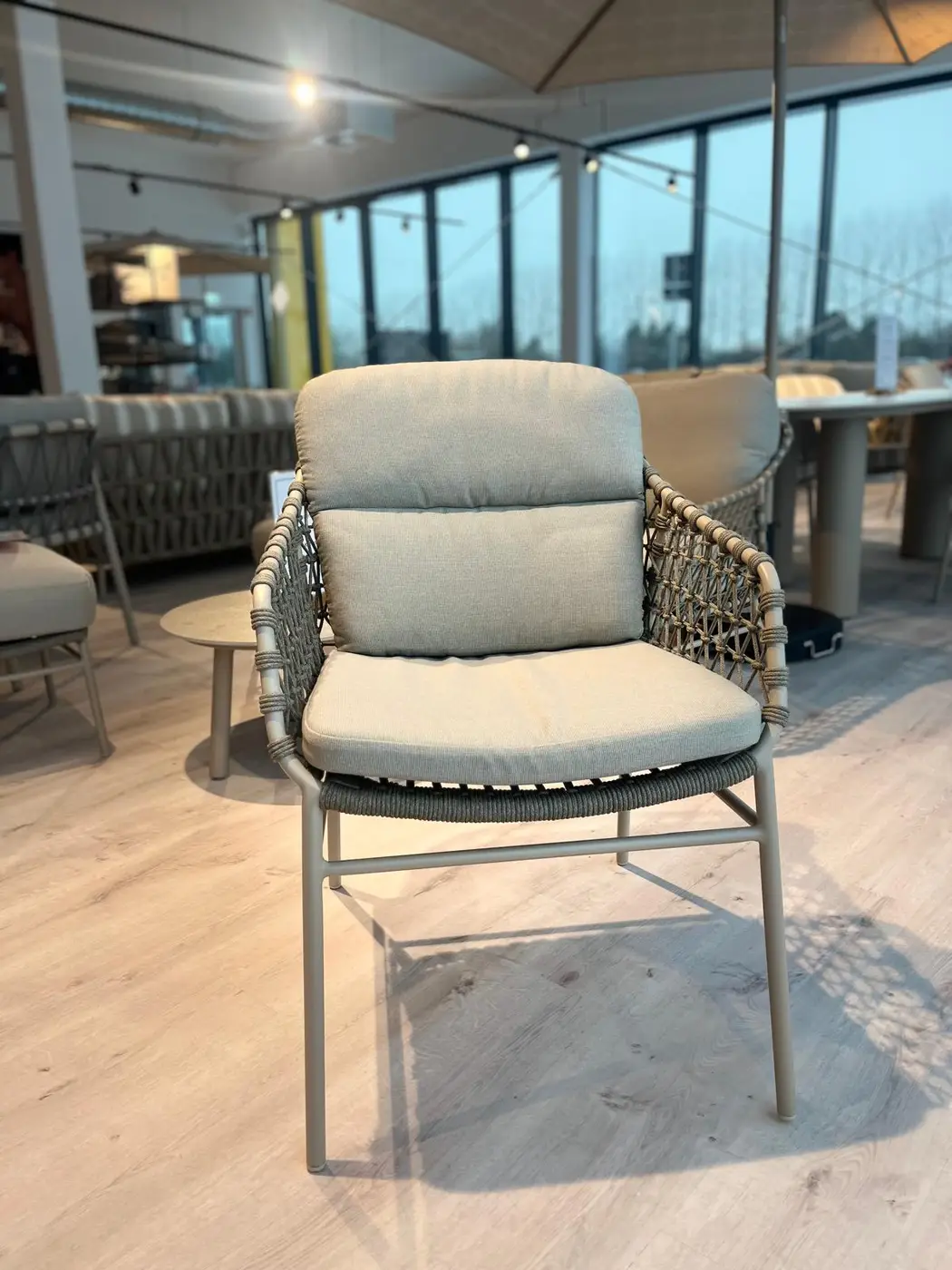 Der Kos Dining Stuhl, ein gepolsterter Terrassenstuhl mit geflochtenen Armlehnen, wird im Innenbereich auf einem hellen Holzboden gezeigt.