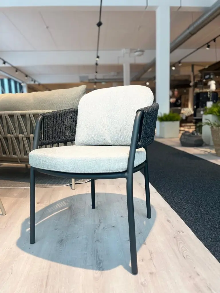 Der Pinto Dining Stuhl hat ein schwarzes Gestell, geflochtene Seiten und ein helles Kissen, das in einem Ausstellungsraum gezeigt wird.