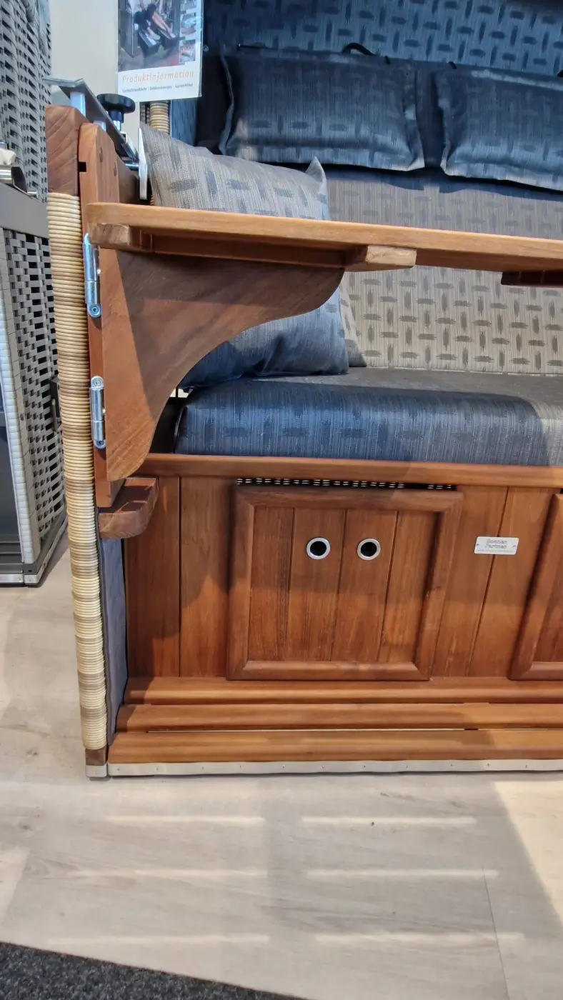 Seitenansicht des Strandkorbs Präsident Teak - Musterabverkauf mit blauen Polstern, Becherhaltern und klappbarem Tablett auf der Armlehne. Gartenmöbel und Grills kaufen in Rostock, Mecklenburg-Vorpommern.